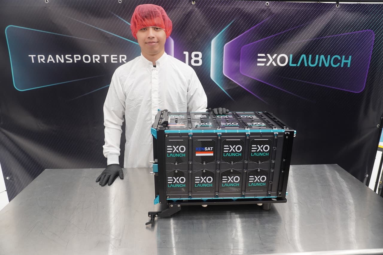 Ken in cleanroom garb beside the EXOLaunch dispenser carrying KENSAT, in front of a Transporter-18 banner